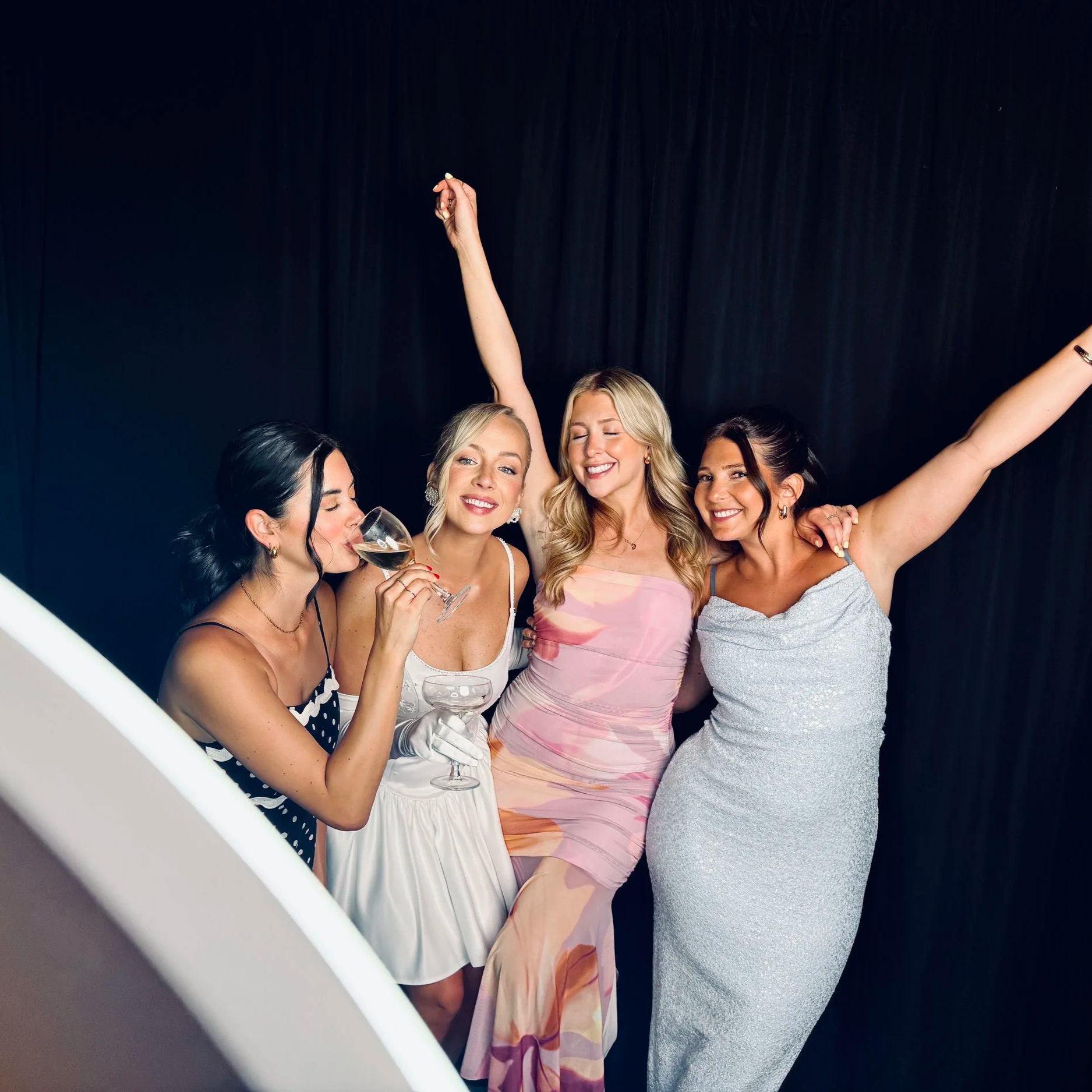 Four women celebrating at a party, with one drinking wine and others smiling and dancing against a black background.