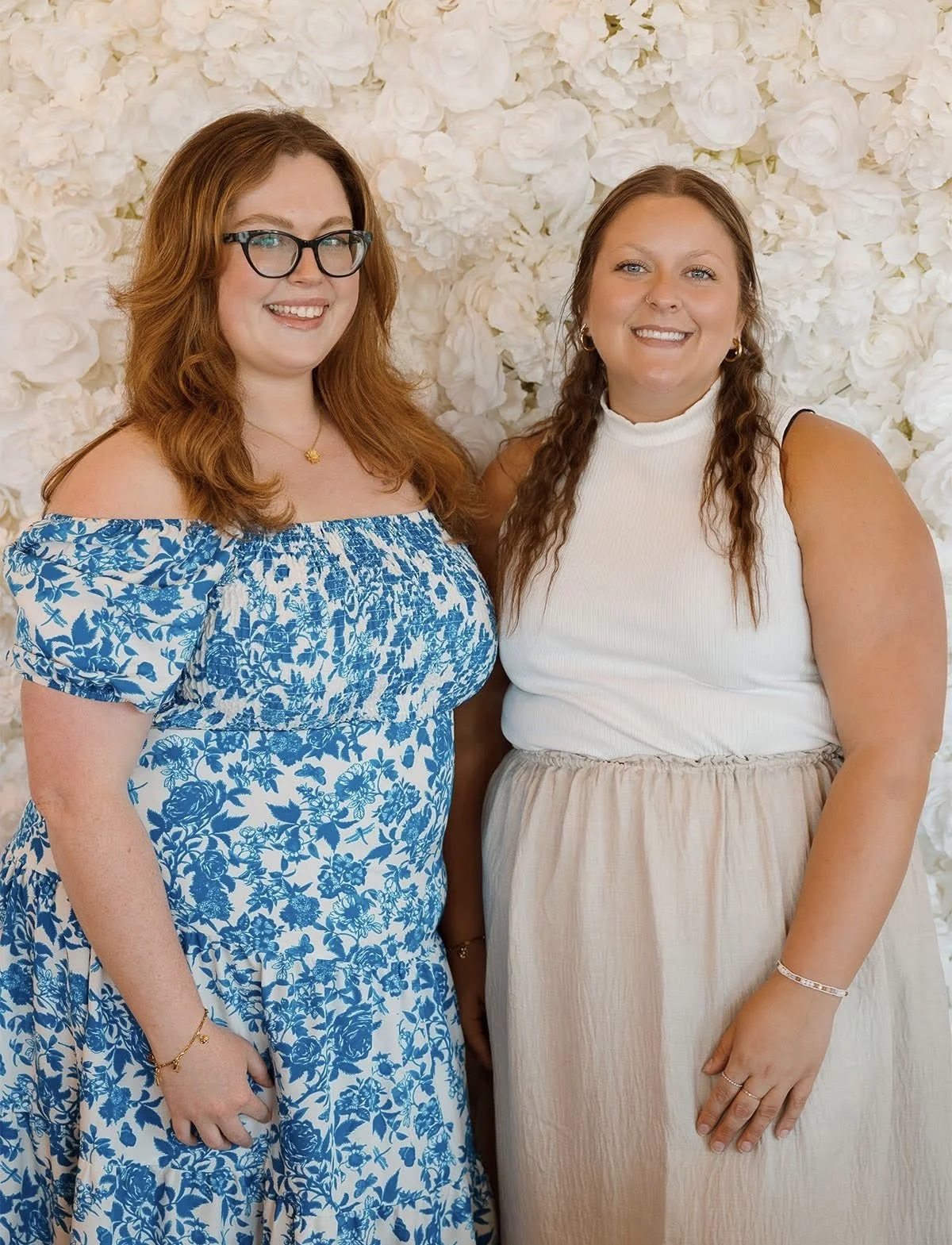 Two women standing in front of a white floral backdrop, smiling at the camera.