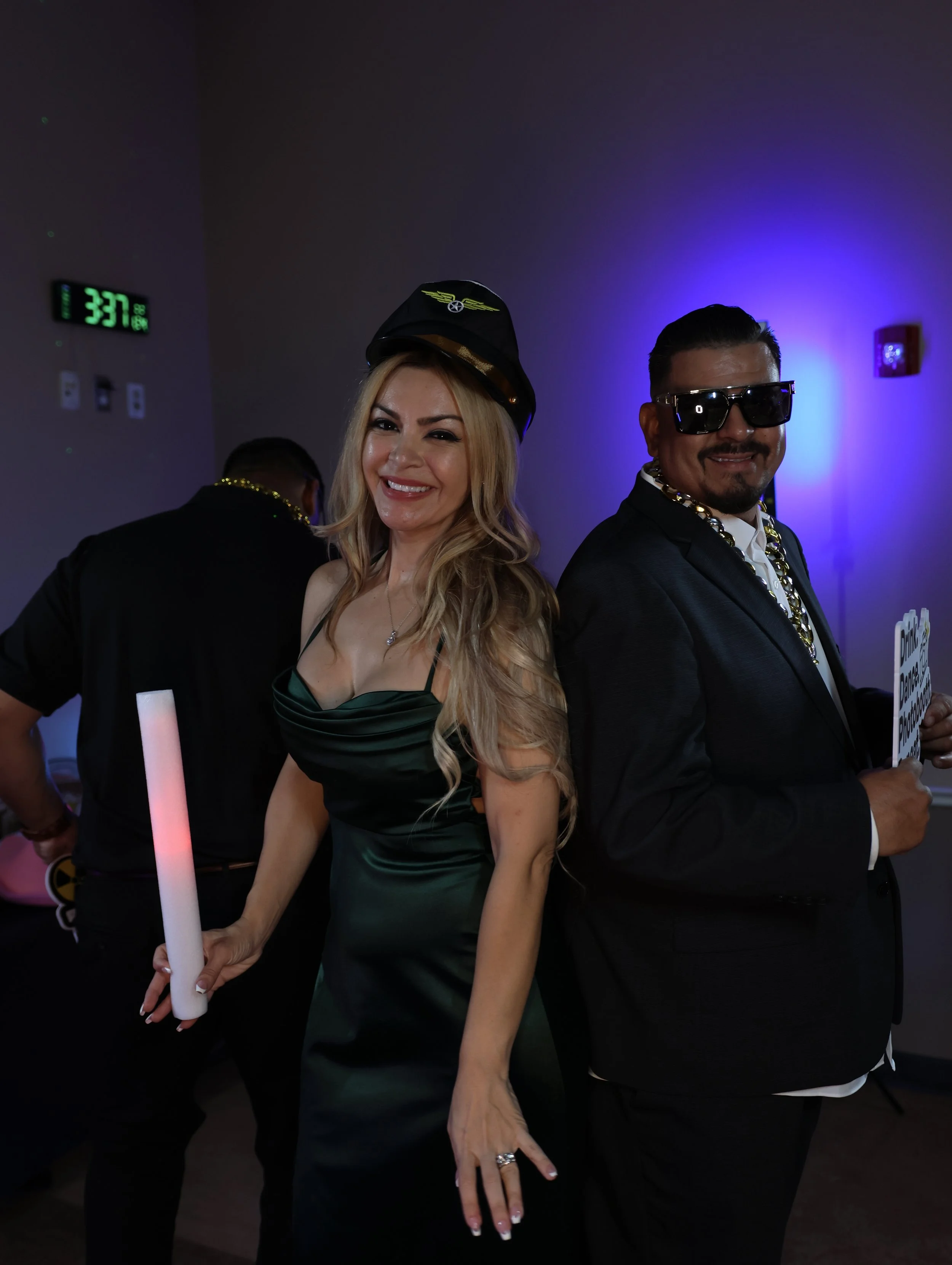 Two people dressed in party attire, a woman with long blonde hair in a dark green satin dress and a man with short dark hair in a black suit, both wearing costume accessories and holding glow sticks, posing at a lively indoor party with colorful ligh
