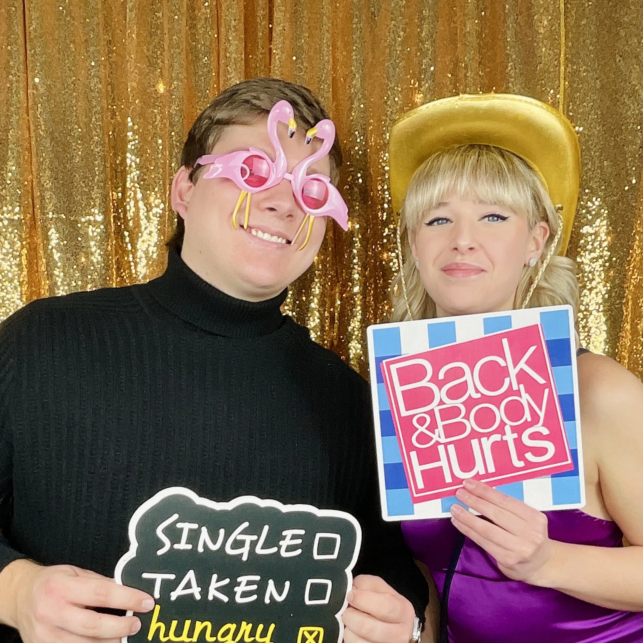 Two people at a photo booth with a gold sequin backdrop. The person on the left is wearing a black turtleneck and pink flamingo-shaped sunglasses, holding a sign that reads "SINGLE O TAKEN" with one check box marked. The person on the right, dressed 