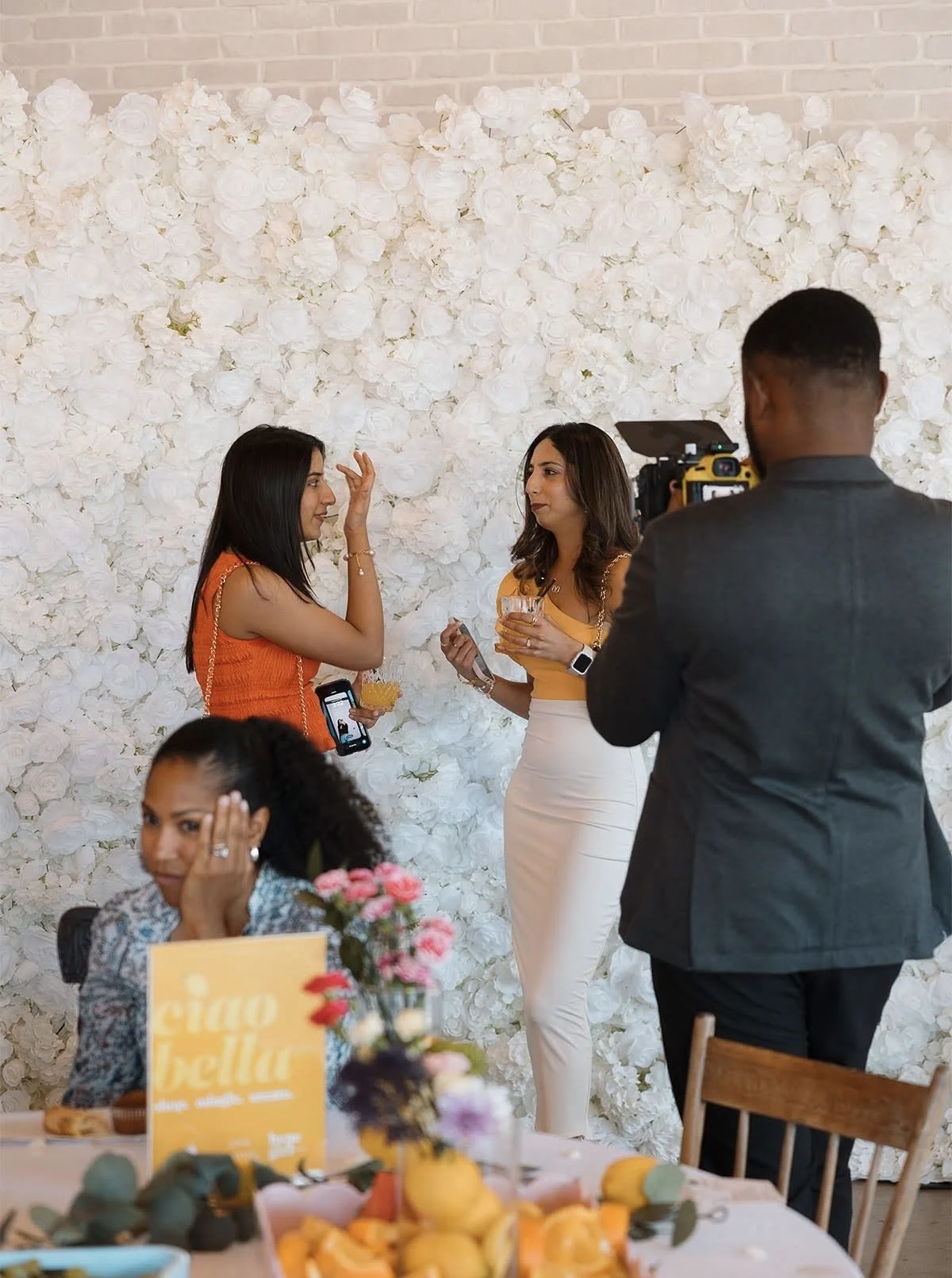 Two women are talking during a social event, with a floral white backdrop. One woman in an orange top holding a drink, the other in a yellow top and white pants, also holding a drink. A man in a gray suit stands with his back to the camera, filming t
