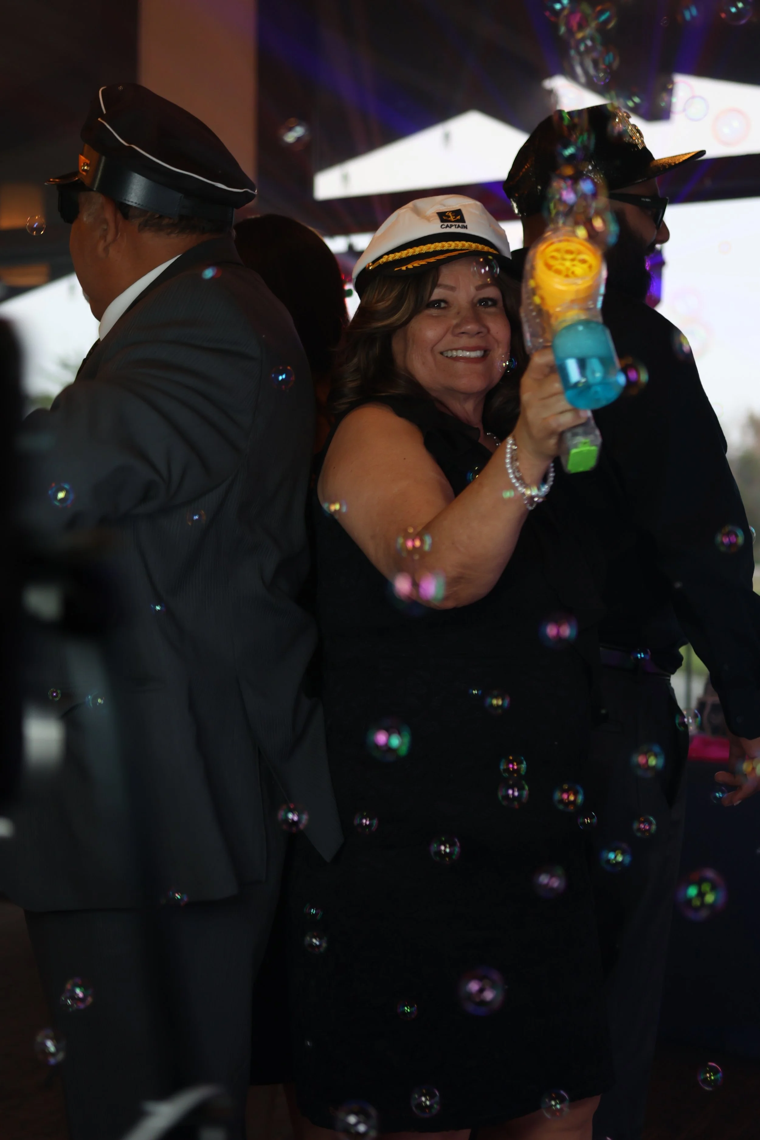 A smiling woman in a black dress with a white captain's hat, holding a bubble gun, surrounded by bubbles at a celebration or party.