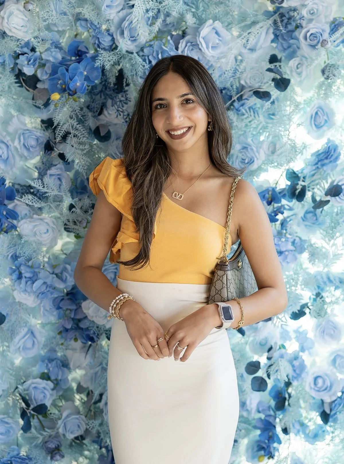 A woman with long dark hair wearing a yellow one-shoulder top and white skirt, standing in front of a blue floral backdrop, smiling at the camera.