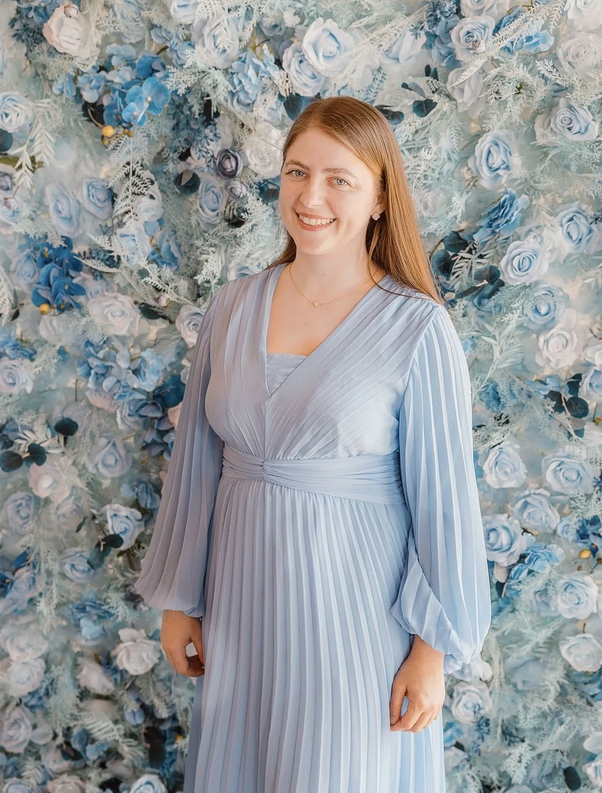 A woman with long red hair smiling, wearing a light blue pleated dress in front of a decorative wall made of blue and white roses and foliage.