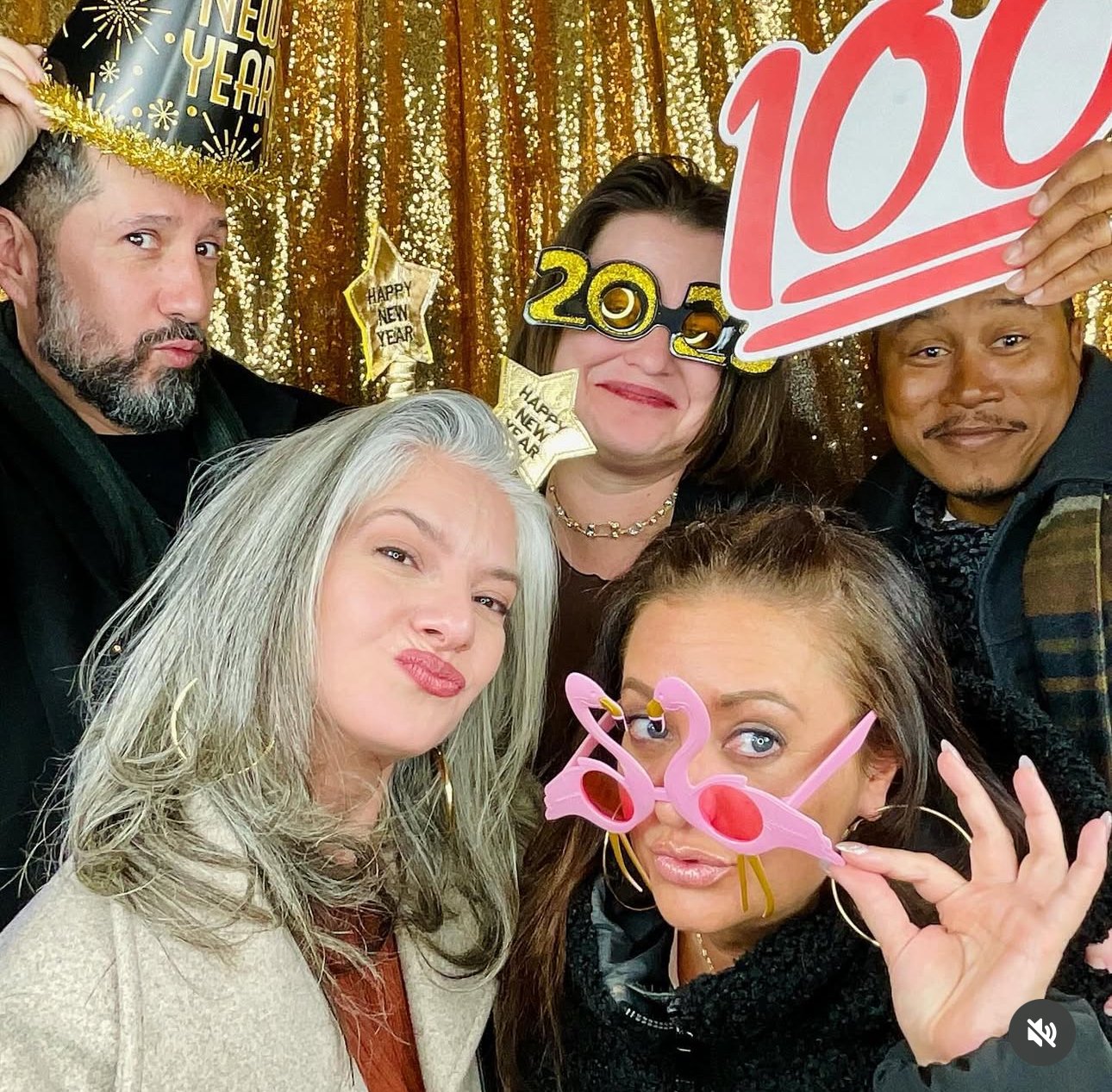 Group of five people celebrating New Year's Eve, wearing party accessories and colorful glasses, with gold sequin backdrop and New Year decorations.