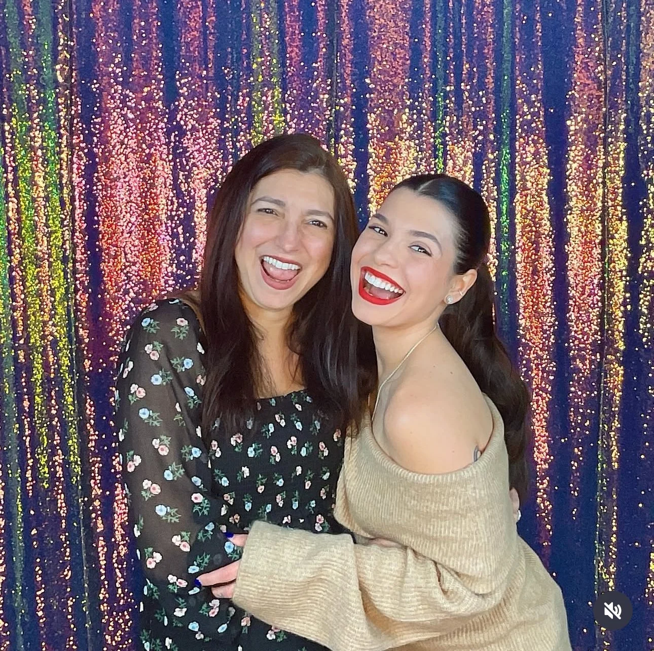 Two women smiling and laughing in front of a colorful, shimmering background, one with dark brown hair wearing a black floral dress, and the other with black hair in a ponytail wearing a beige off-shoulder top.