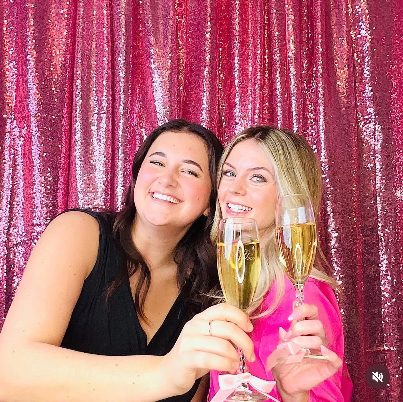 Two women smiling and holding glasses of champagne in front of a pink, sequined curtain background.