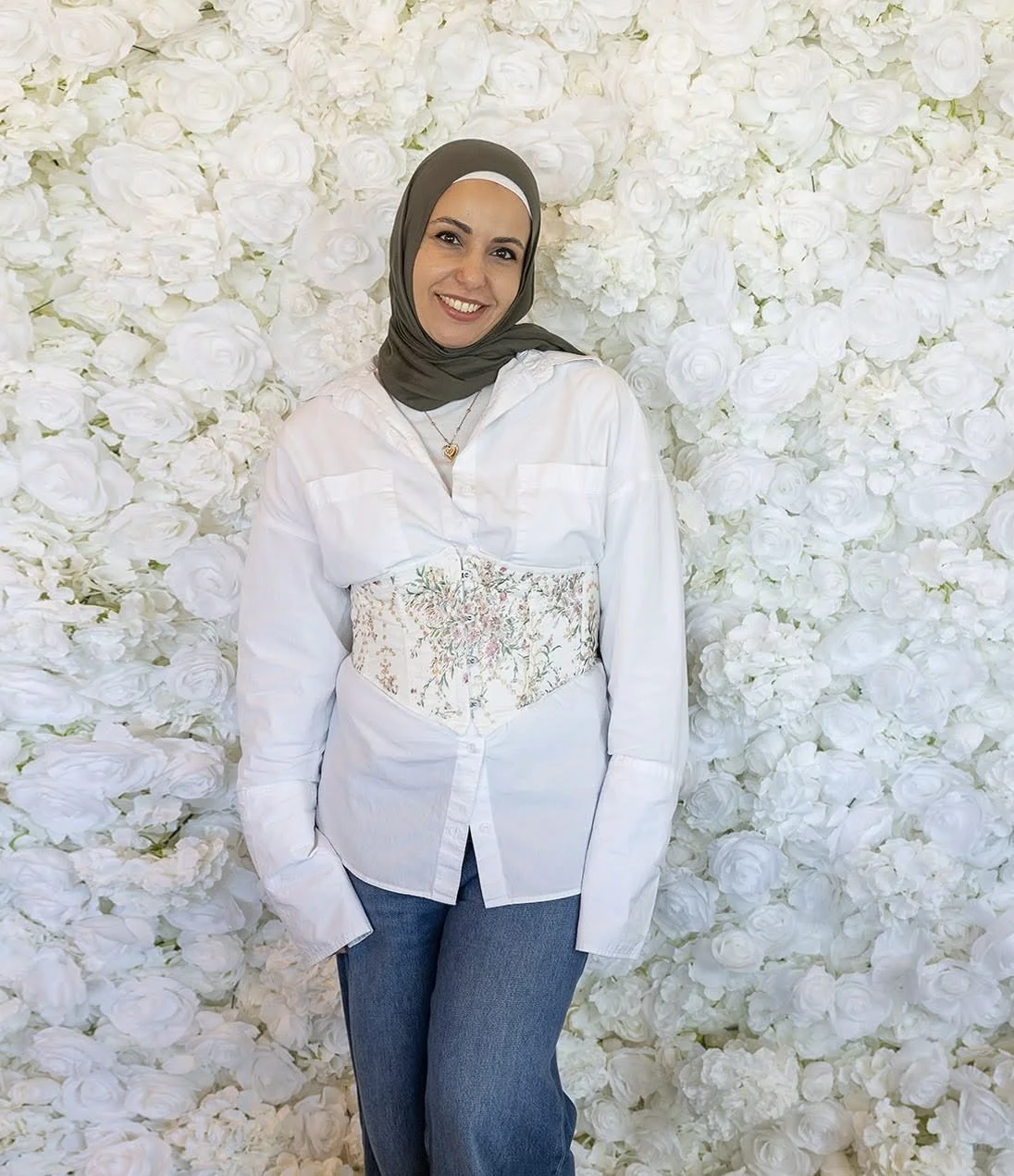 A woman smiling in front of a wall of white roses, wearing a gray hijab, white shirt with floral embroidery, and blue jeans.
