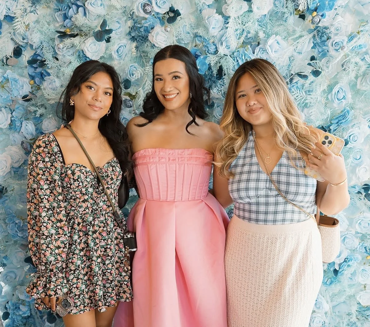 Three women standing in front of a blue floral backdrop, smiling at the camera, dressed in colorful outfits.