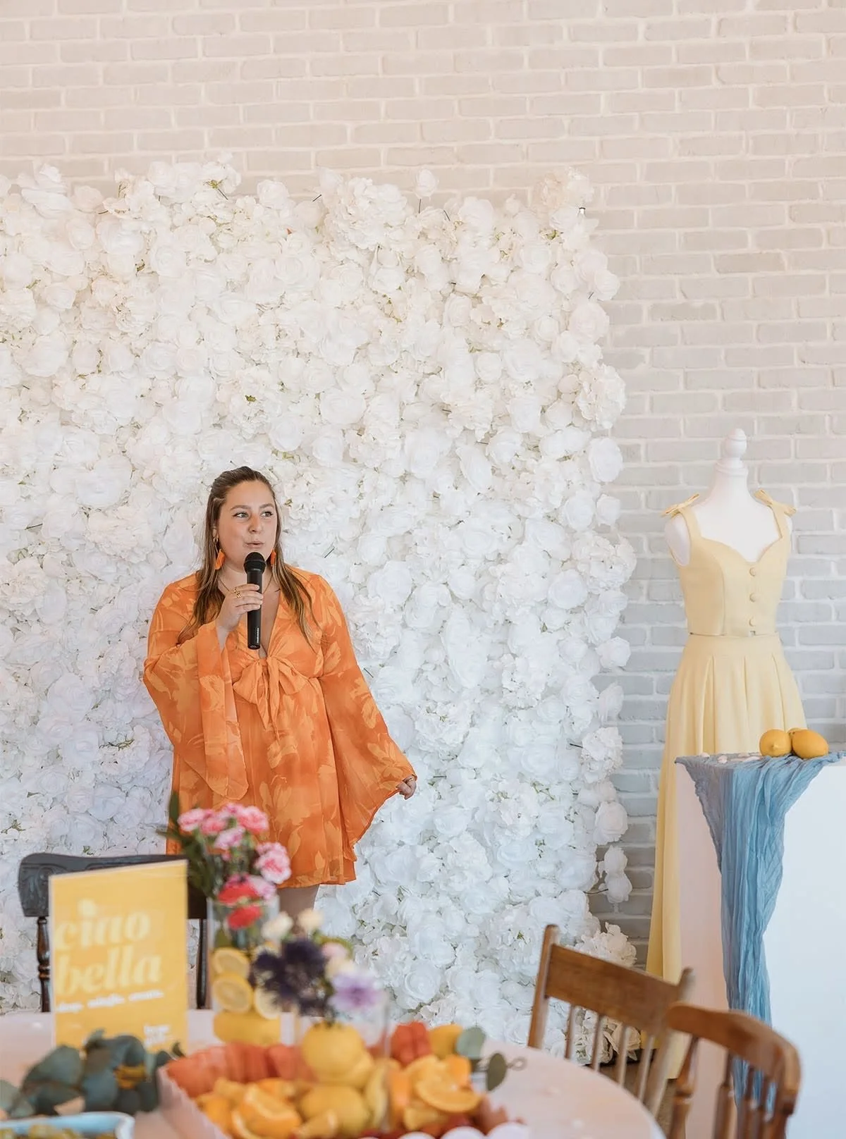 A woman in an orange dress holding a microphone, standing in front of a white floral backdrop at a decorated event space with a white brick wall and a mannequin dressed in a yellow dress.