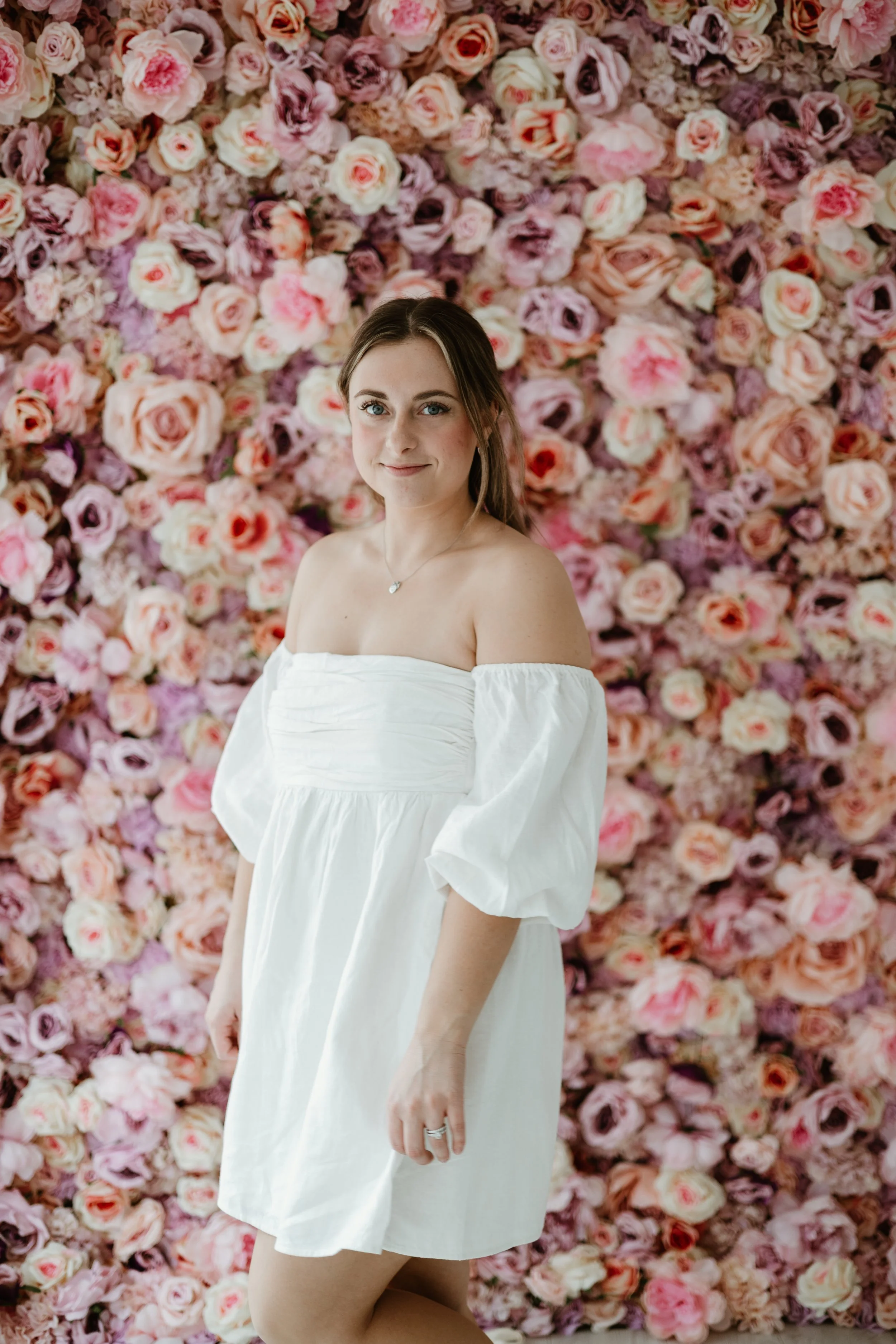 Young woman in white off-shoulder dress standing in front of a wall covered with pink, peach, and purple roses.