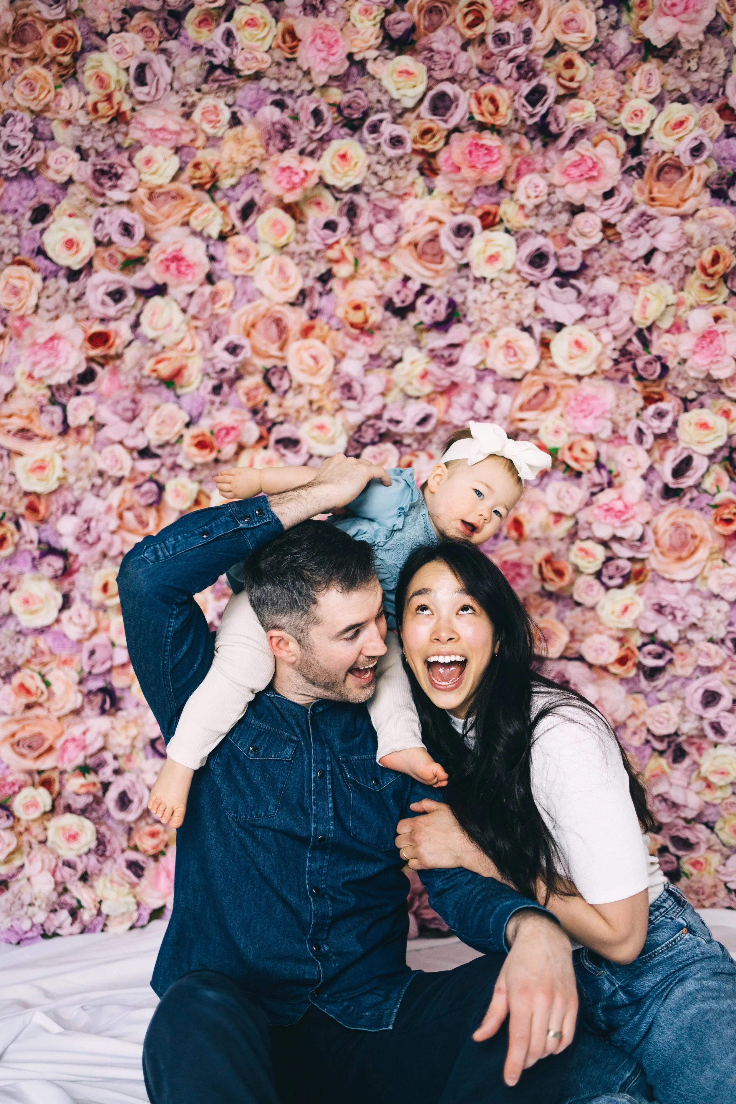 A happy family of three enjoying a moment together, sitting in front of a pink and purple flower wall, with the father holding the young girl on his shoulders and the mother smiling brightly.