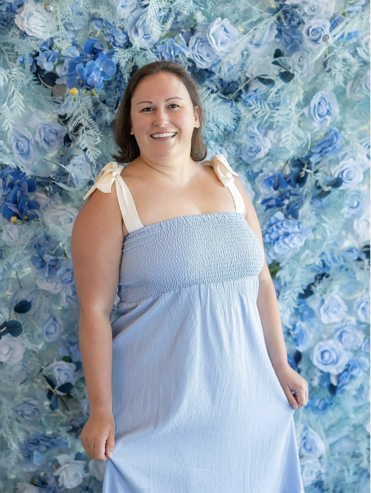 Smiling woman in a light blue dress standing in front of a blue floral backdrop.