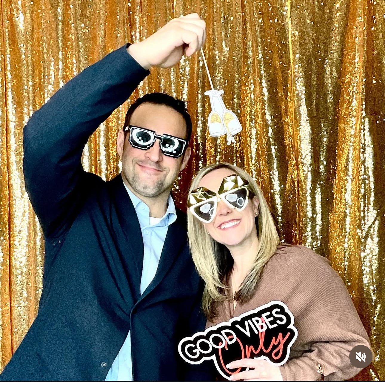 A man and woman are smiling and posing for a photo with a gold sequin backdrop. The man is holding a paper cutout of two bottles hanging from a string above his head. Both are wearing large, decorative sunglasses, and the woman is holding a sign that