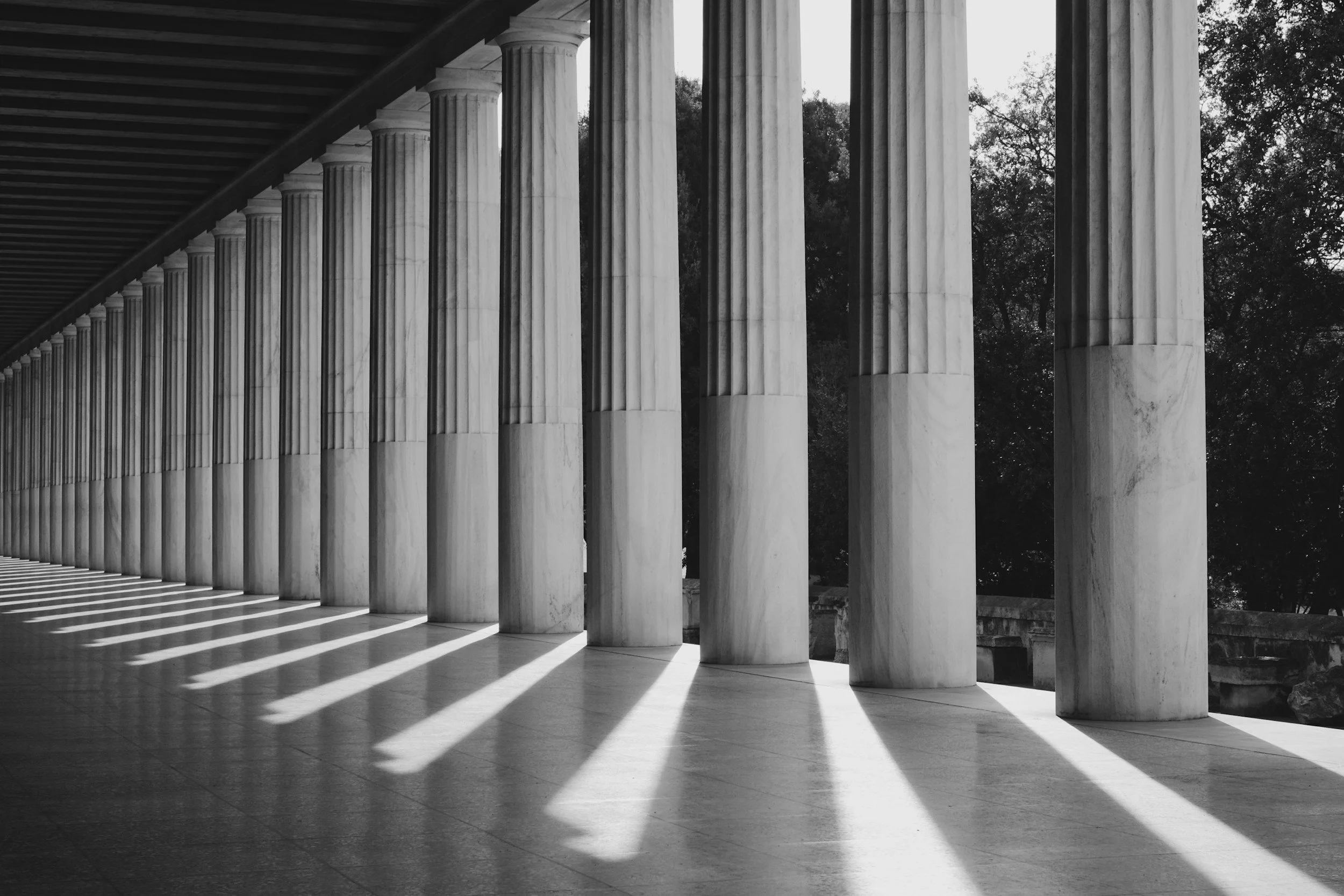 Black and white photo of a row of marble columns casting shadows on the ground, in front of a building with classical architecture.
