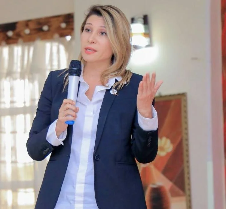 A woman in a navy blazer and white shirt holding a microphone and speaking or presenting indoors.