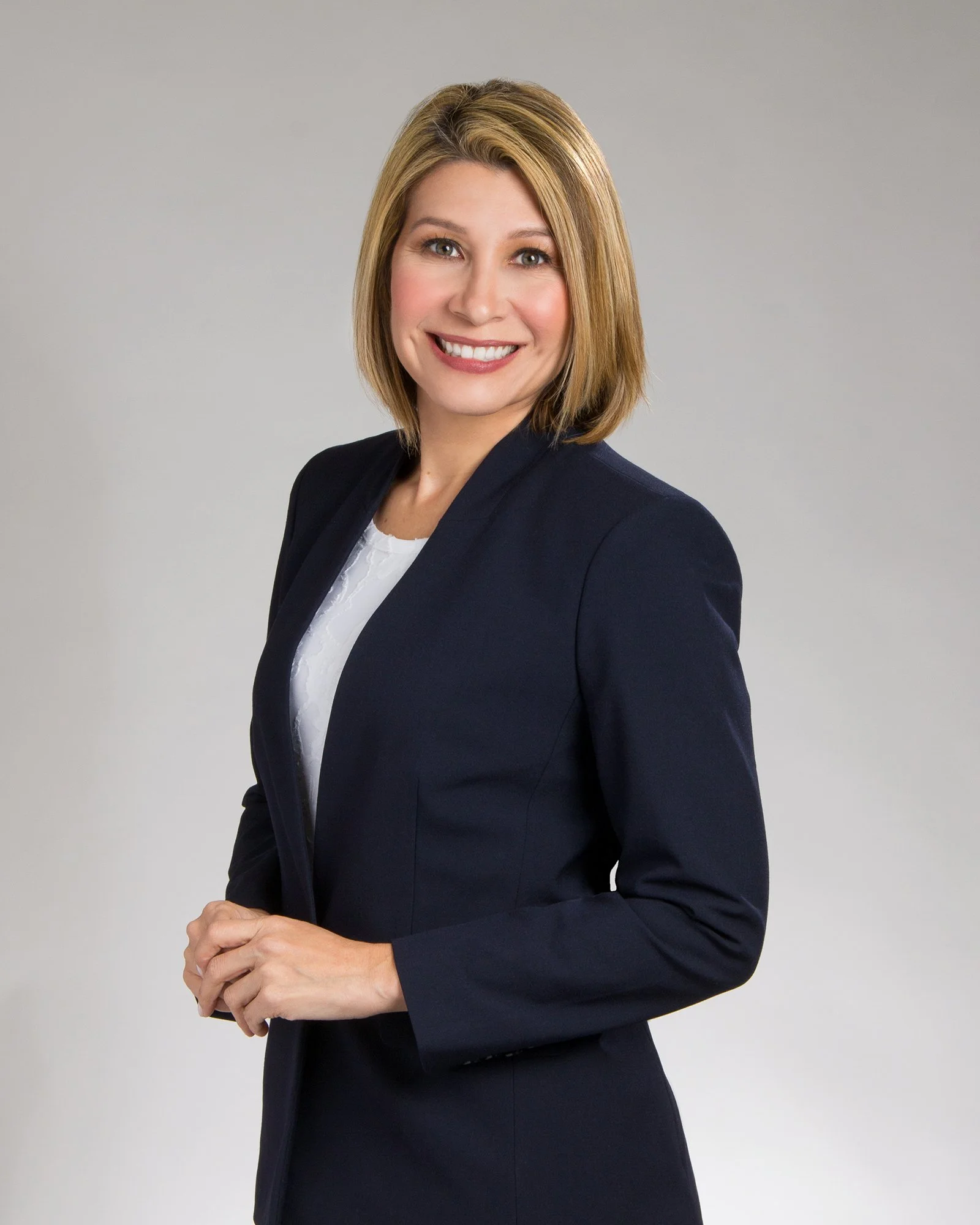 A woman with shoulder-length blonde hair smiling, wearing a navy blazer over a white blouse, standing against a plain grey background.