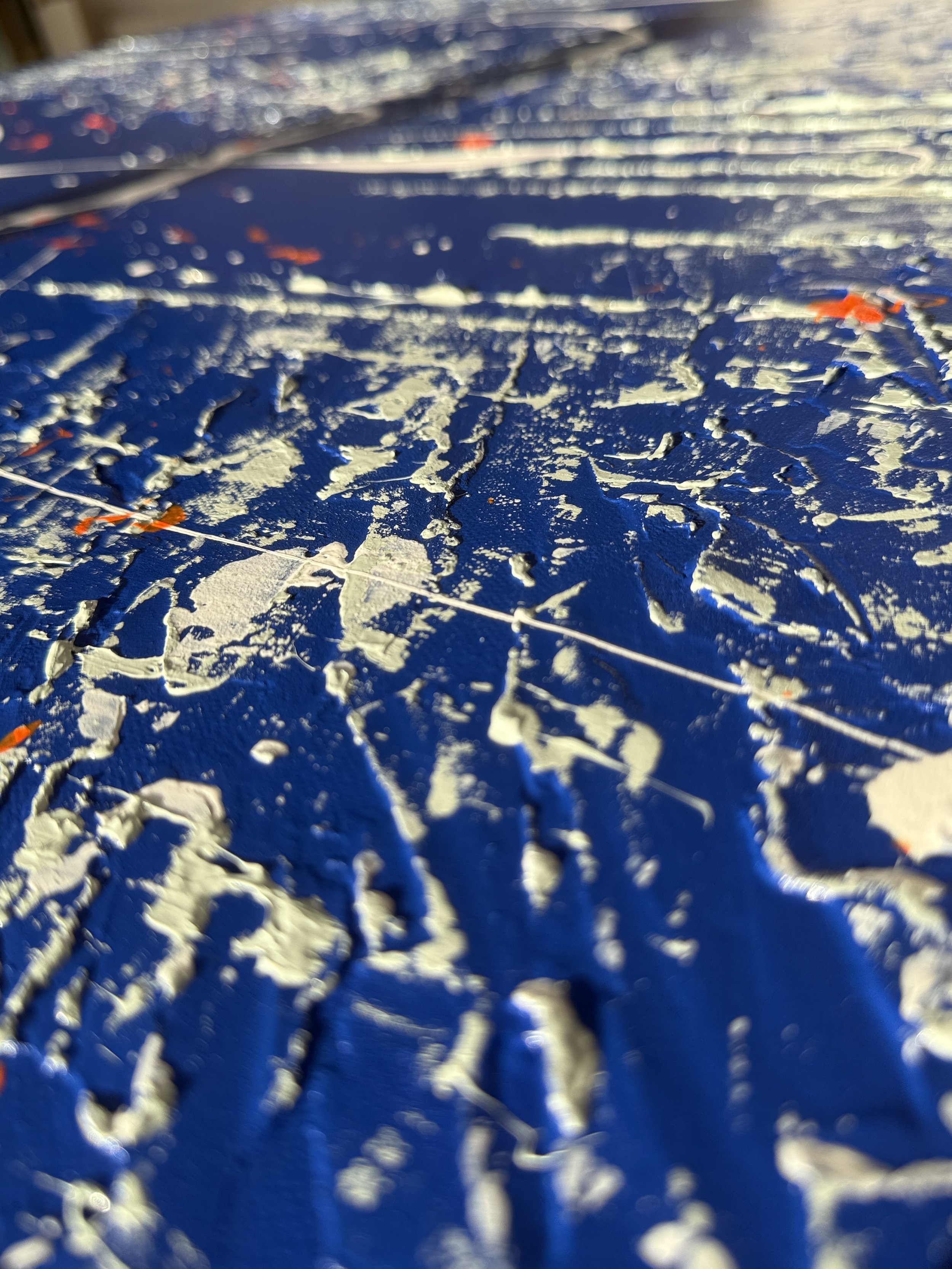 Close-up d'une surface bleue avec des traces épaisses de peinture beige ou blanche, avec quelques petites touches d'orange, vue en angle étalée et texturée.
