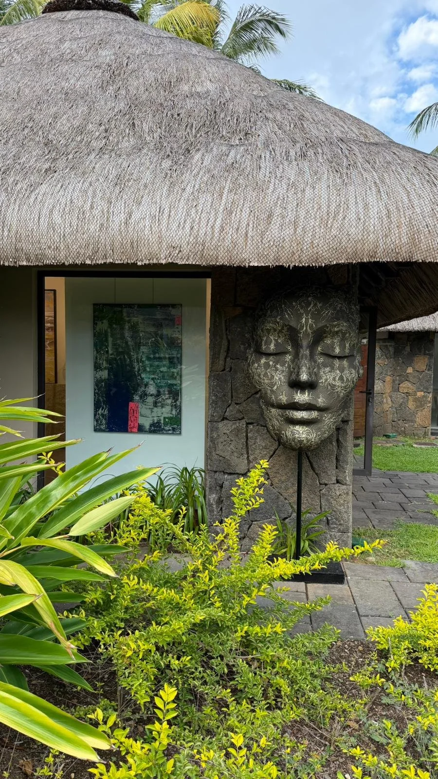 Une sculpture de visage féminin stylisé en métal fixé sur un mur en pierre, entourée de verdure tropicale, sous un toit en chaume.