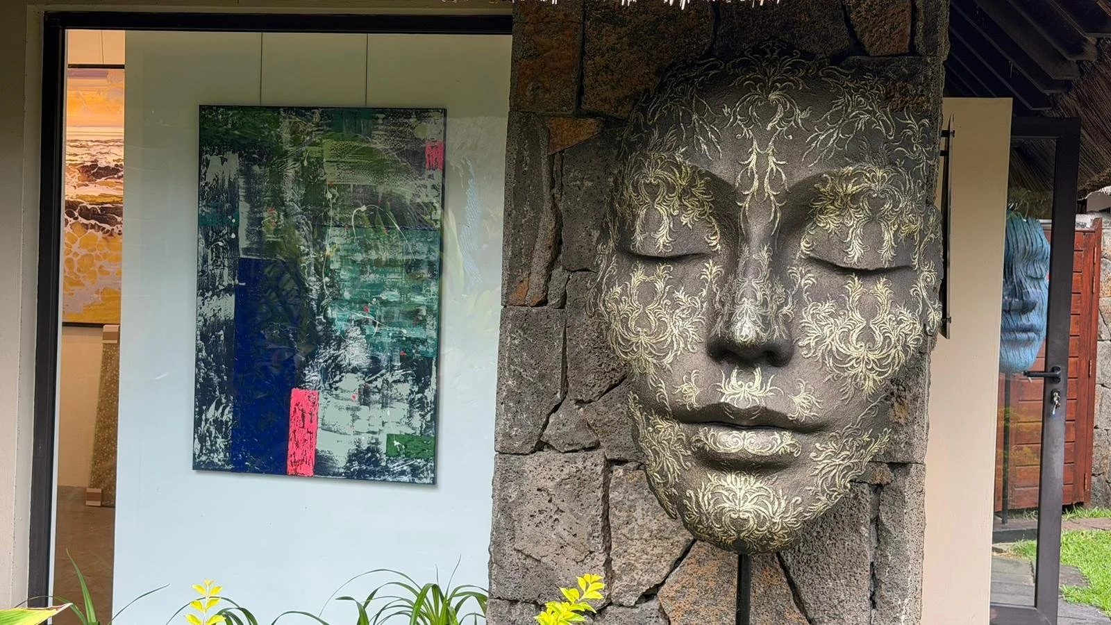 Une sculpture murale en forme de visage humain en pierre avec des détails de motifs floraux et dorés. La sculpture est fixée sur un mur en pierre, à côté d'une pièce d'art moderne avec des couleurs vives et des motifs abstraits.