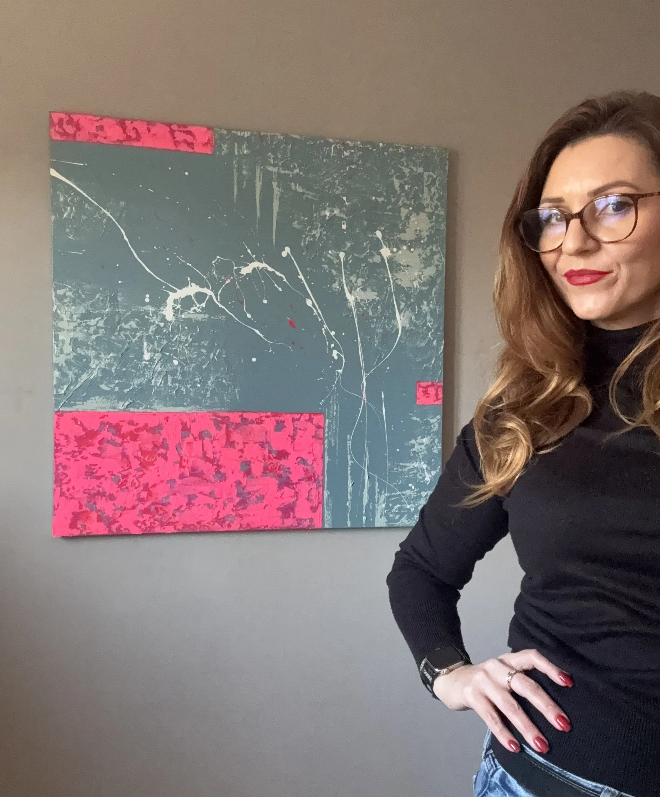 Une femme avec des cheveux bruns, porte des lunettes, une chemise noire et un jean, pose devant une peinture abstraite avec des couleurs gris, rose et blanc.