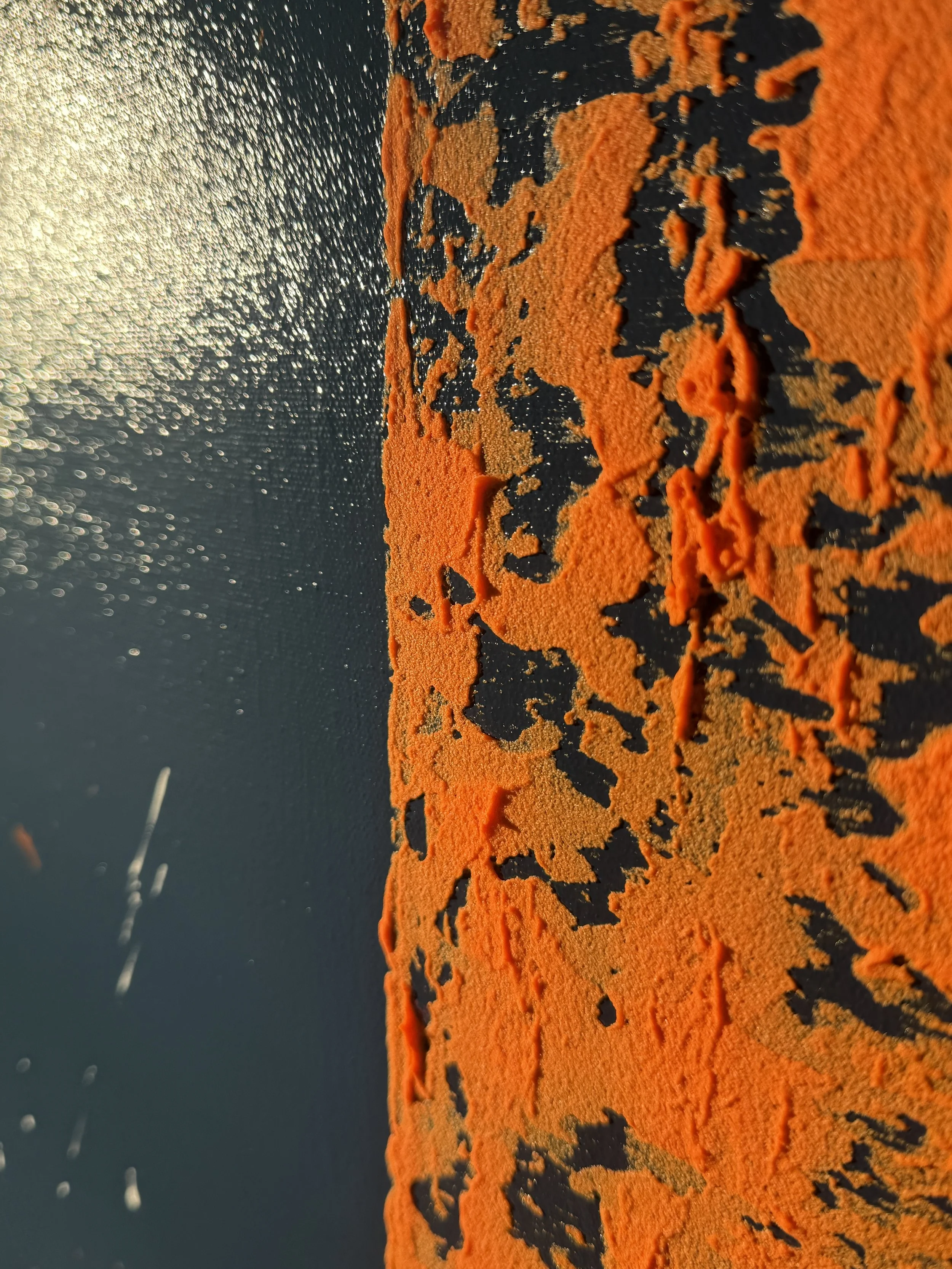 Surface rocheuse orange et noire, avec des textures rugueuses, au premier plan, la lumière du soleil se reflète sur une surface d'eau derrière.