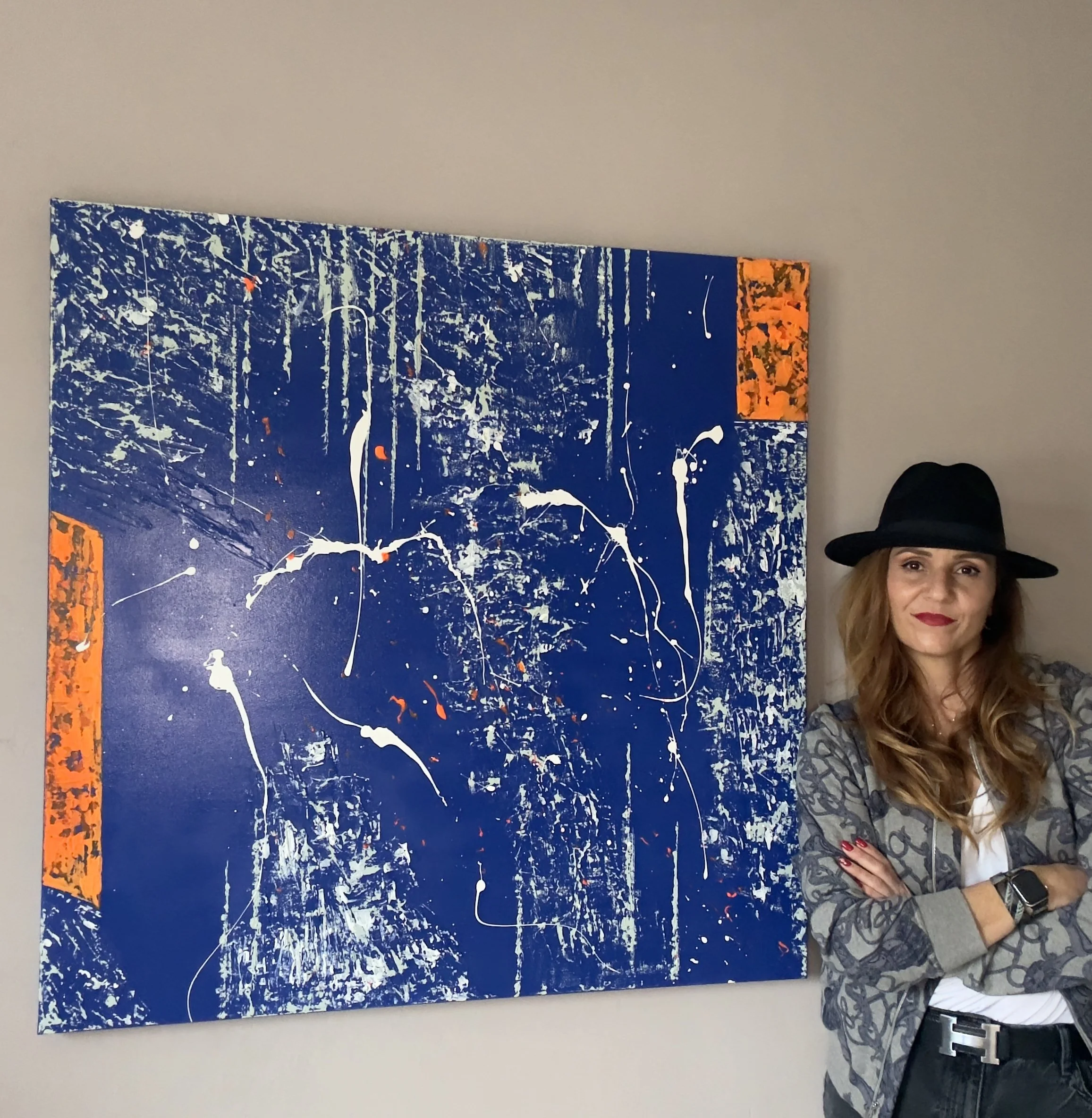 Femme portant un fedora noir, vêtue d'une chemise grise à motifs et d'un jean, se tenant à côté d'une œuvre d'art abstraite sur un mur beige.