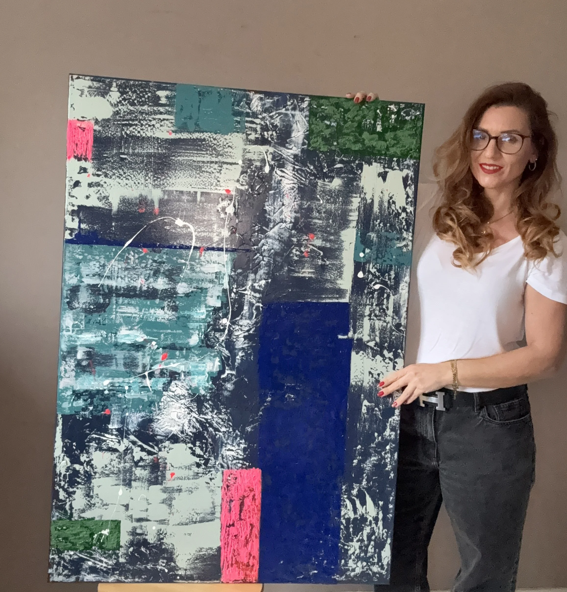 Une femme aux cheveux roux bouclés, portant des lunettes, tient une peinture abstraite complexe avec des couleurs vives et des textures variées.