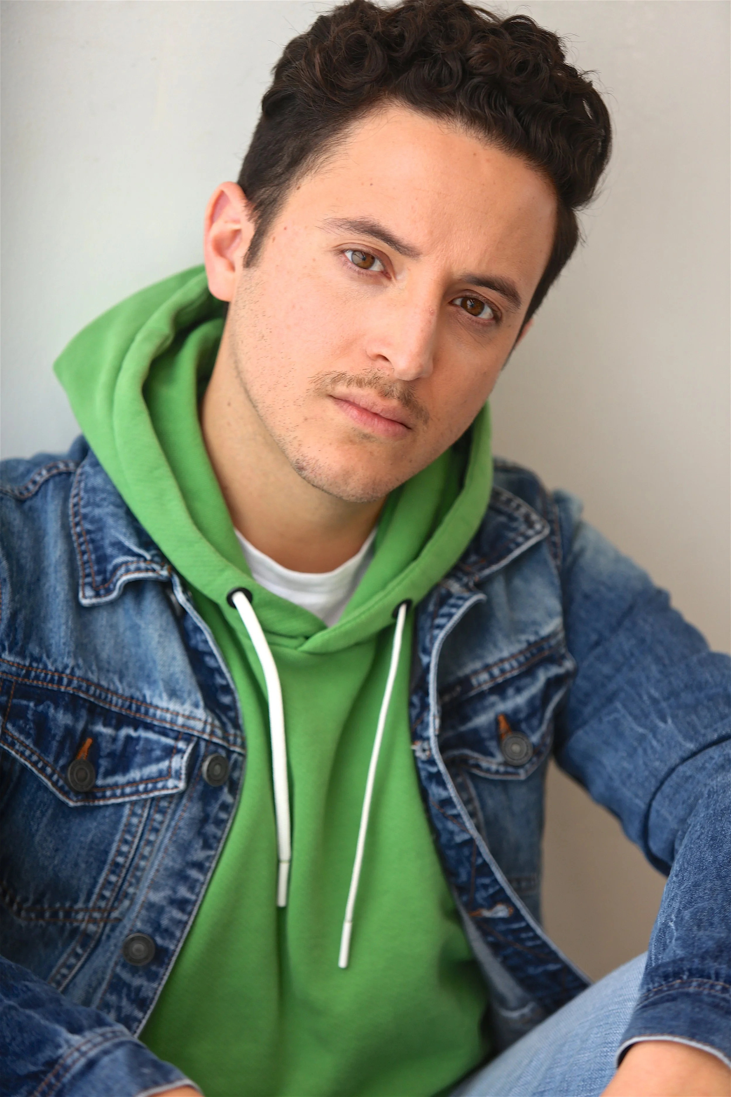 Professional portrait of Alex de la Torre, a bilingual actor, creator, and storyteller based in Los Angeles, wearing a green hoodie and denim jacket and looking directly at the camera.
