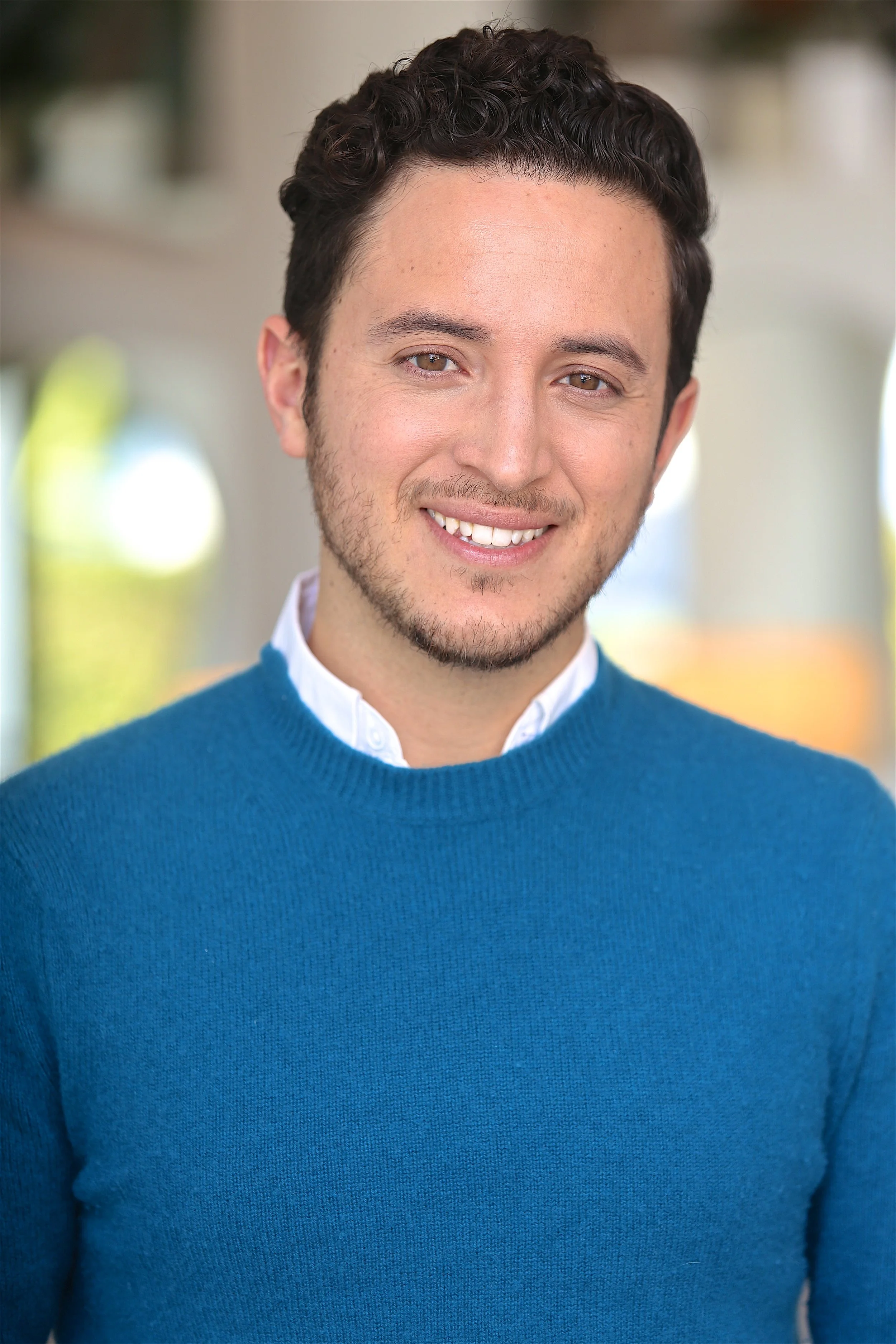 Professional portrait of Alex de la Torre smiling while wearing a blue sweater and glasses. Alex is a bilingual actor, creator, and storyteller based in Los Angeles, pictured in a friendly and approachable pose.
