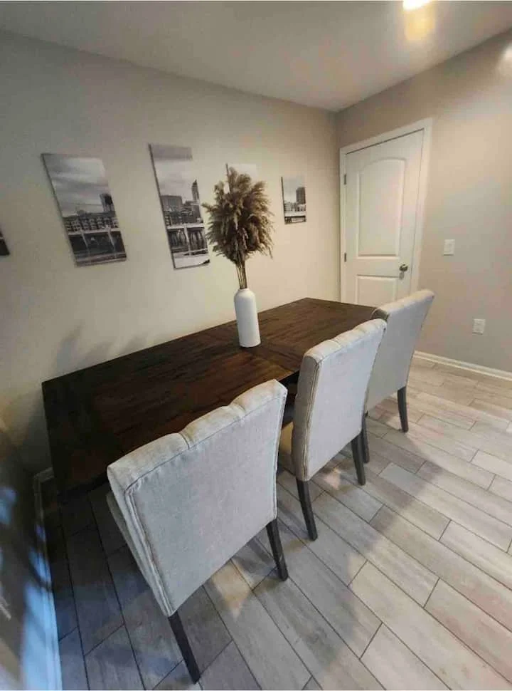 A dining room with a wooden table, three beige upholstered chairs, a white vase with dried plants, and black-and-white cityscape artwork on the wall.