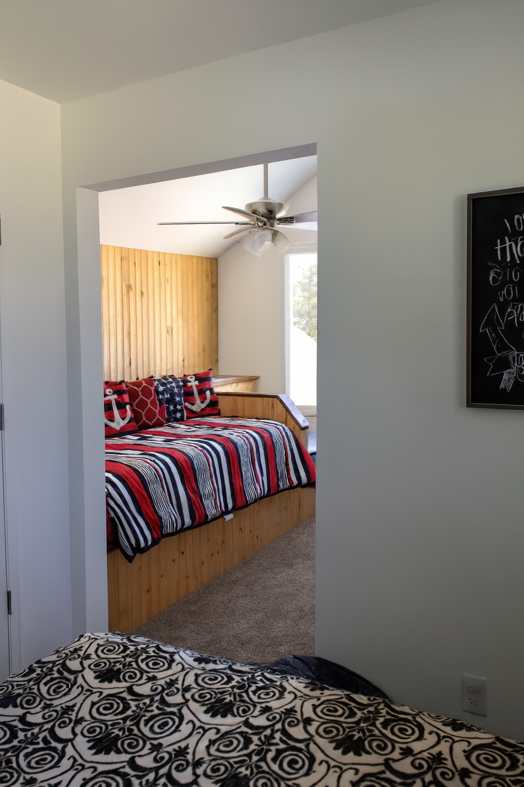 A cozy bedroom with a motivational blackboard on the wall, a bed with black and white patterned bedding, and a view into an adjoining room featuring a wooden bed with patriotic-themed bedding and decorative pillows with anchors and stars.