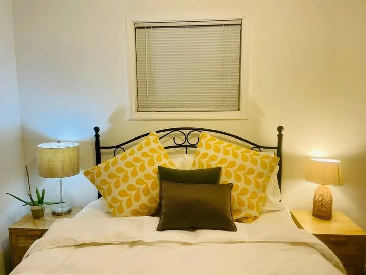 A neatly made bed with yellow and white heart-patterned pillows and a dark accent pillow, flanked by two wooden nightstands with lamps, one with a small potted plant, in front of a window with closed blinds in a bedroom.