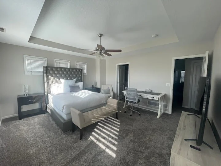 A spacious bedroom with a large bed, tufted headboard, and two pillows. There are bedside tables on each side, one with a lamp. A cushioned bench sits at the foot of the bed. The room has gray walls, a ceiling fan, and three small windows above the b
