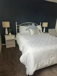 A bedroom with a white bed, matching white nightstands with lamps, and a dark blue wall.