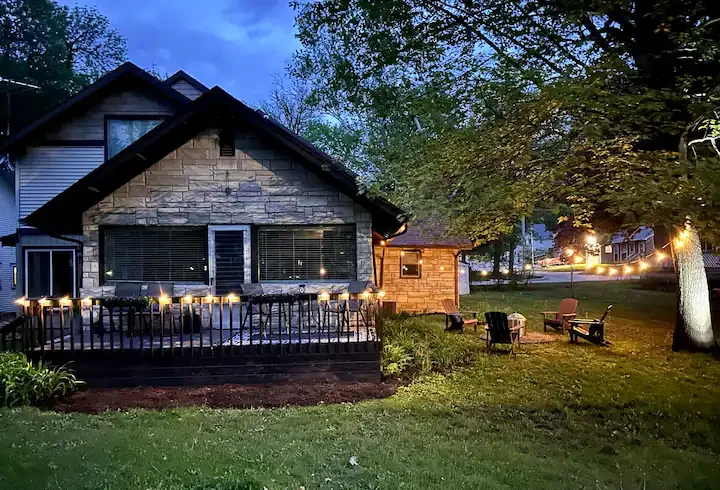 A backyard scene at dusk featuring a house with a stone facade and an elevated wooden deck with string lights. There are several outdoor chairs and a small fire pit on the lawn, with trees and neighboring houses in the background illuminated by stree