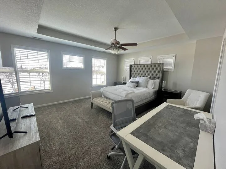 Bedroom with four windows, a large bed with a tall tufted headboard, a small bench at the foot of the bed, a white armchair, a desk with a chair, and a TV on a stand.