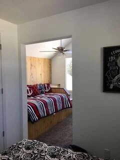 View of a bedroom with a bed featuring a striped red, white, and blue blanket and multiple pillows, visible through a doorway from an adjacent room with white walls.