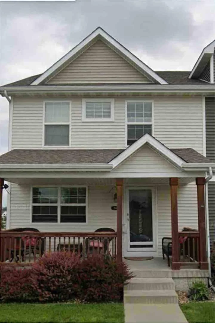 Front view of a two-story house with beige siding, multiple windows, a small front porch with a wooden railing, and steps leading up to the front door, with some shrubs and a green lawn in the foreground.
