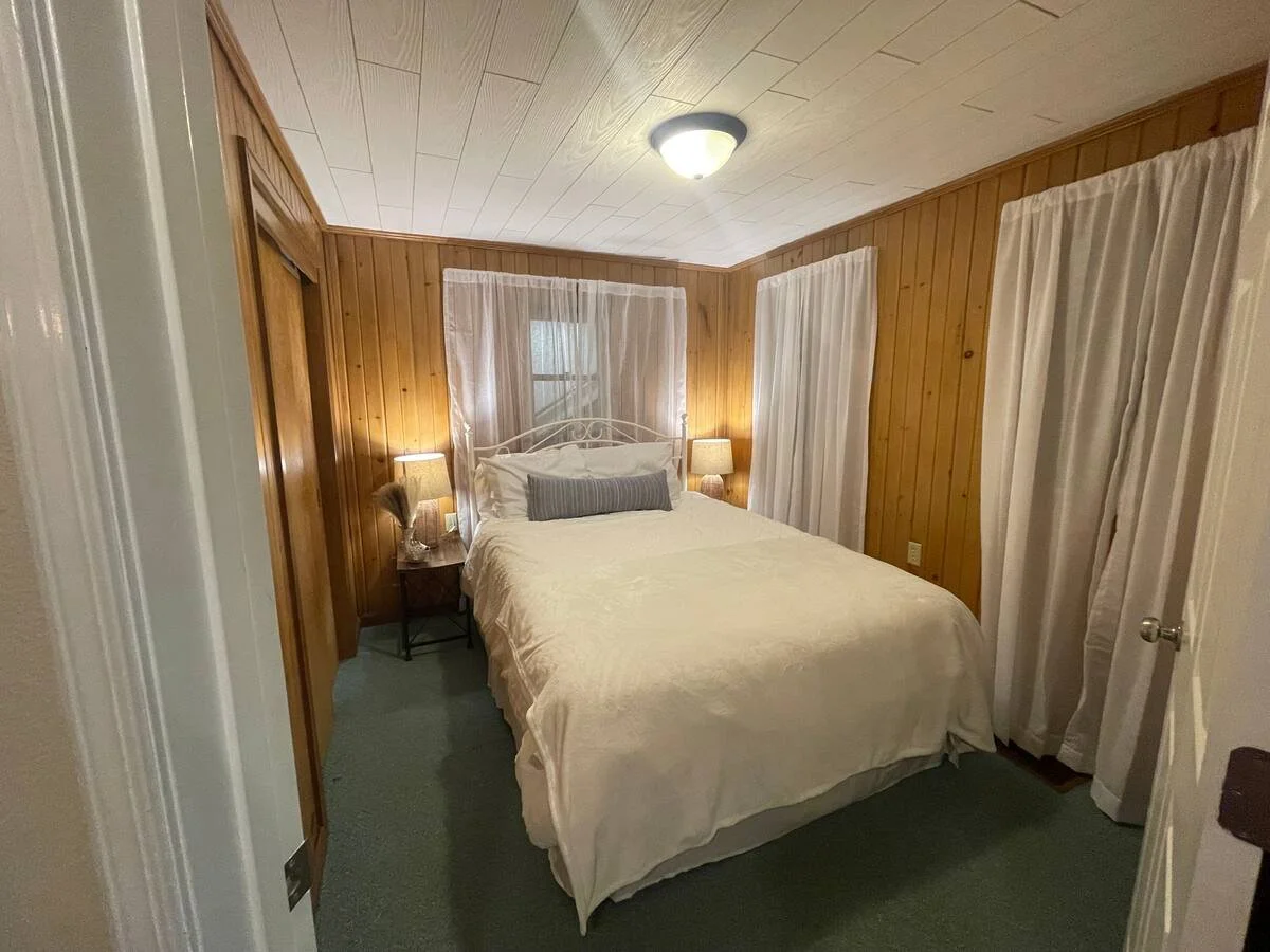 A cozy bedroom with wooden paneled walls, a bed with white bedding, two bedside tables with lamps, and curtains on the windows.