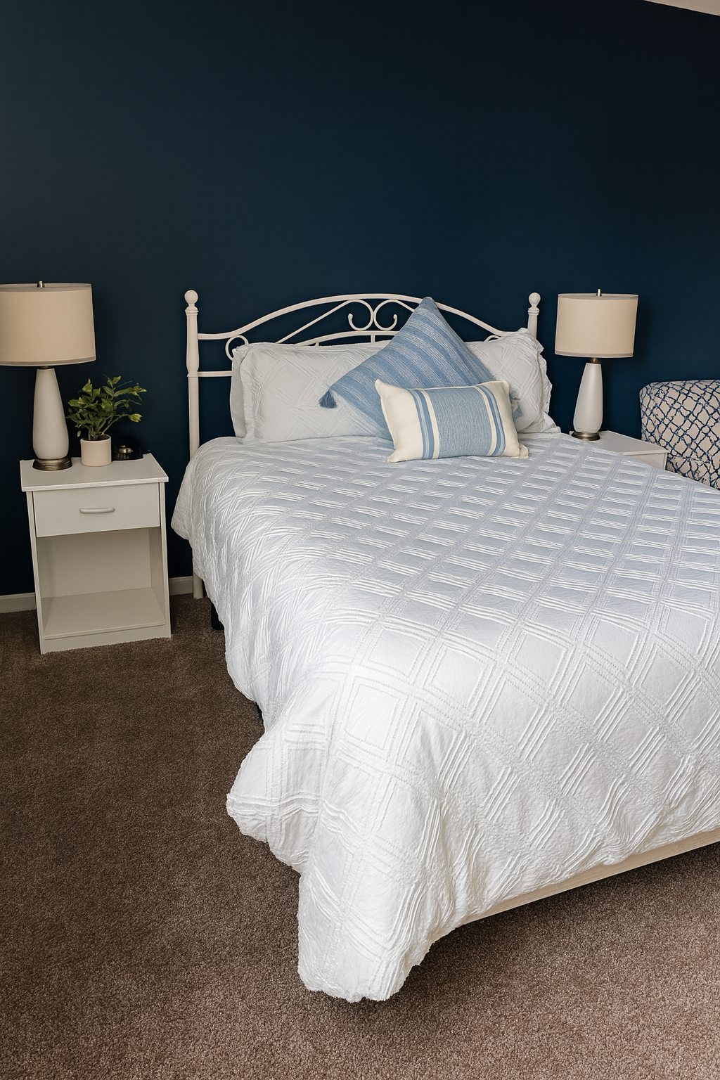A neatly made bed with white bedding and blue decorative pillows, flanked by two white nightstands with lamps, in a bedroom with a dark blue accent wall.