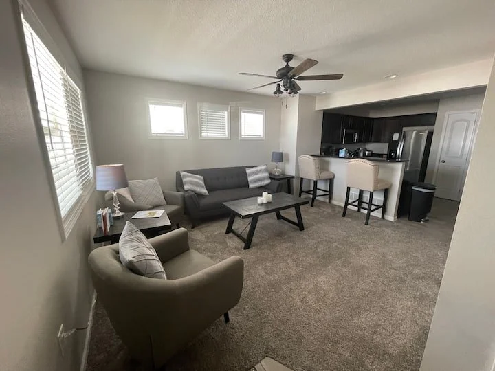 Living room with beige and gray furniture, including a sofa, armchair, and coffee table, with a bar area and barstools in the background.