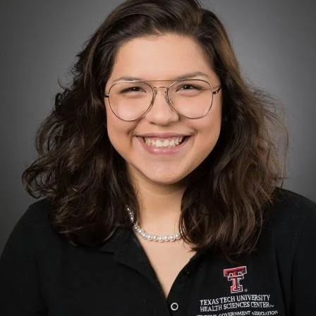 A young woman with brown, wavy hair, wearing glasses, a black shirt with a Texas Tech University logo, and a pearl necklace, smiling against a gray background.