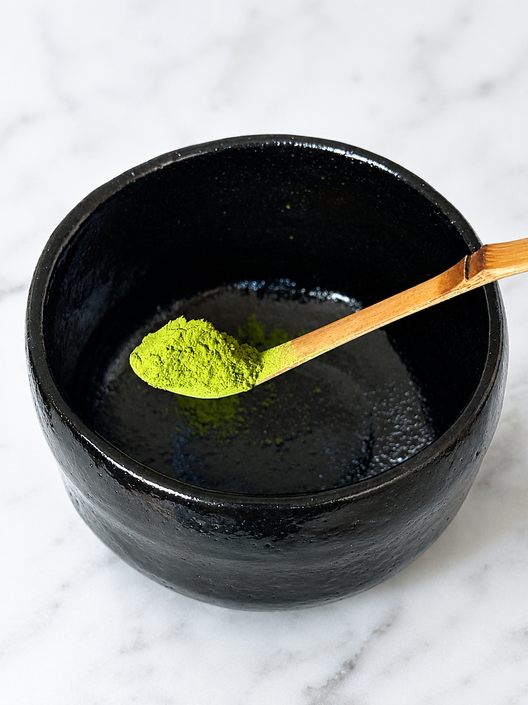 A black bowl with matcha green tea powder on a bamboo spatula and some powder inside, sitting on a white marble surface.