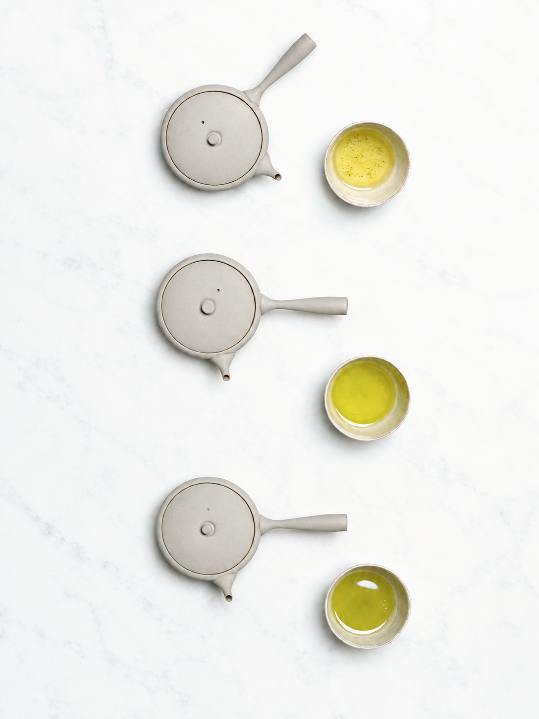 Three teapots with lids and three cups filled with green tea, arranged on a white marble surface.