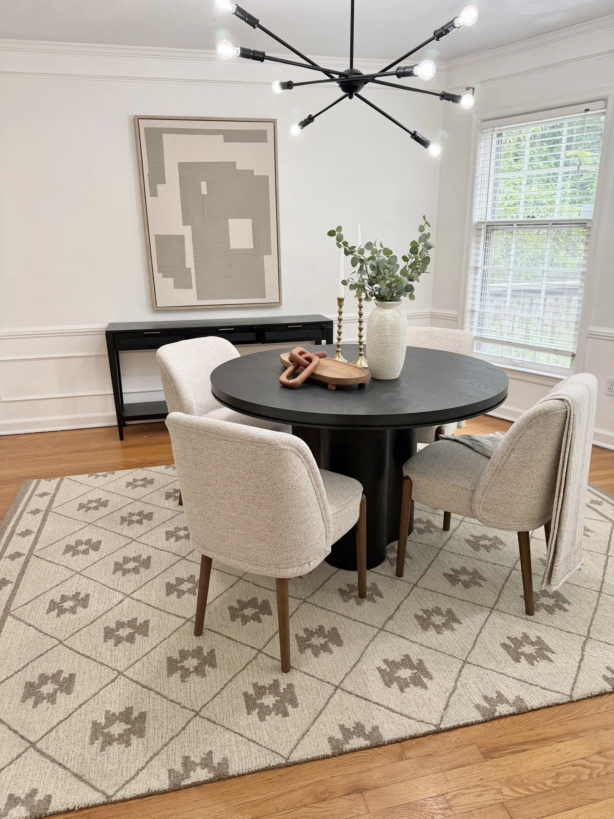 A dining room with a black round table, four beige upholstered chairs, a patterned area rug, a window with blinds, a piece of modern artwork on the wall, a vase with greenery, two gold candlesticks, and wood flooring.