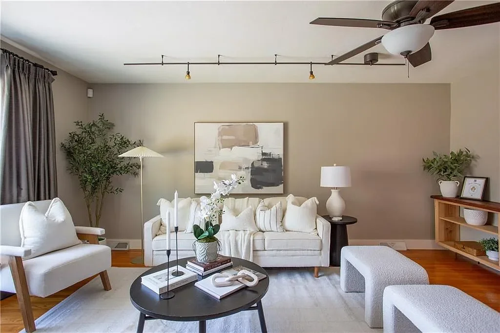 Living room with a white sofa, a white armchair, and a pair of white ottomans. There are decorative pillows, a black coffee table with books and orchids, a lamp, and framed pictures on a wooden shelf. The room has hardwood flooring, a ceiling fan, gr