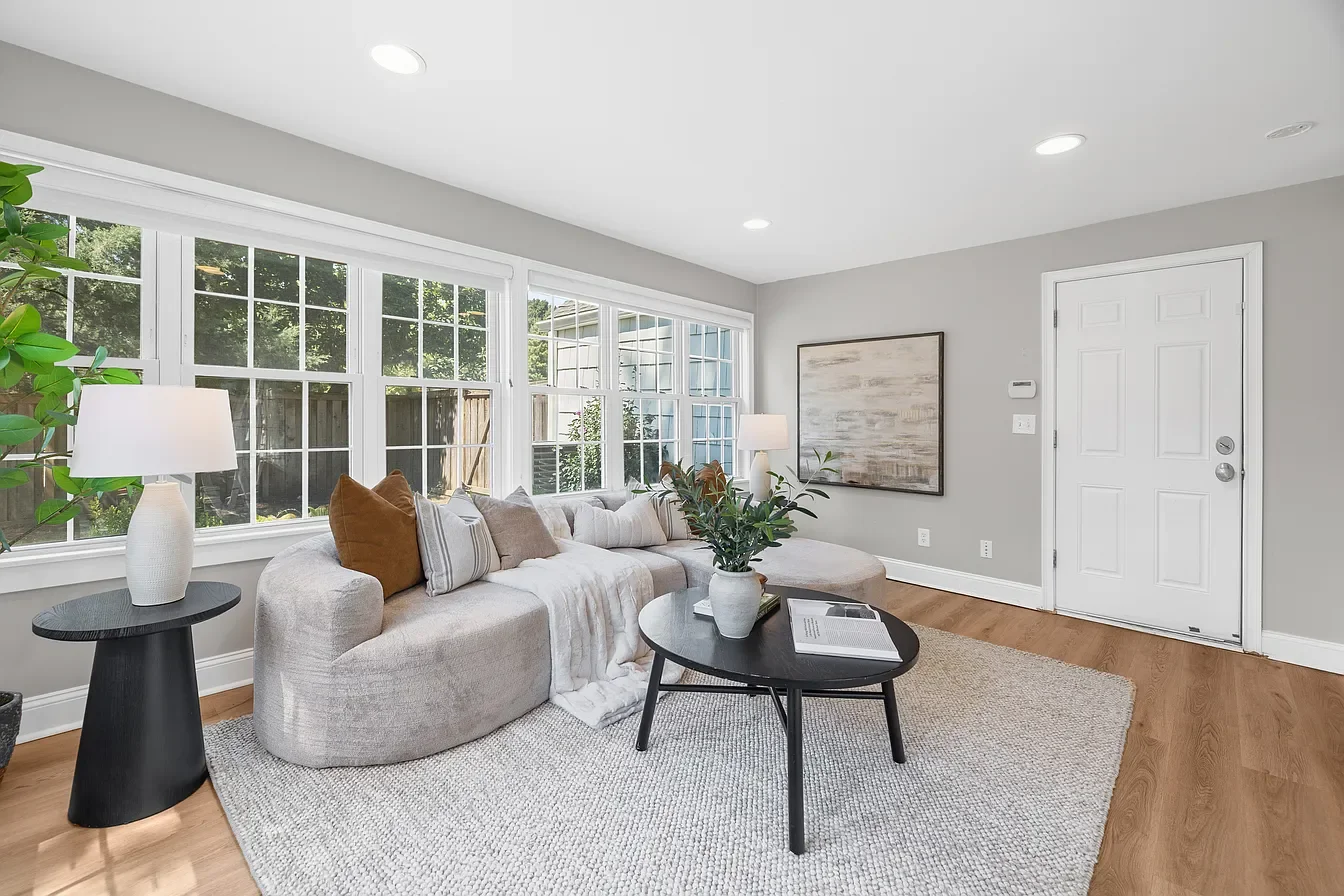 Bright living room with large windows, light-colored sofa with pillows, black coffee table with plant and book, side table with lamp, hardwood floor, and neutral decor.