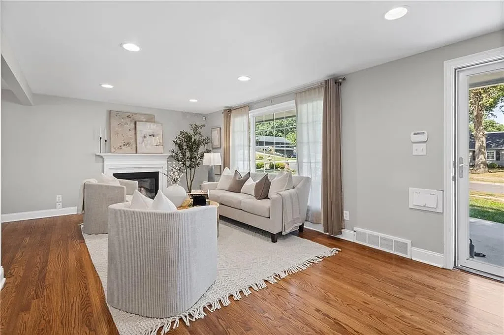 A bright living room with a white sofa, two matching armchairs, a fireplace, and large windows with beige curtains. The room has hardwood floors and a white rug.