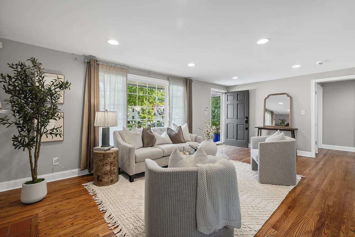Bright living room with light gray walls, hardwood floors, large window with beige curtains, white sofa, two matching armchairs, a beige area rug, floor lamp, decorative mirror, and plants.